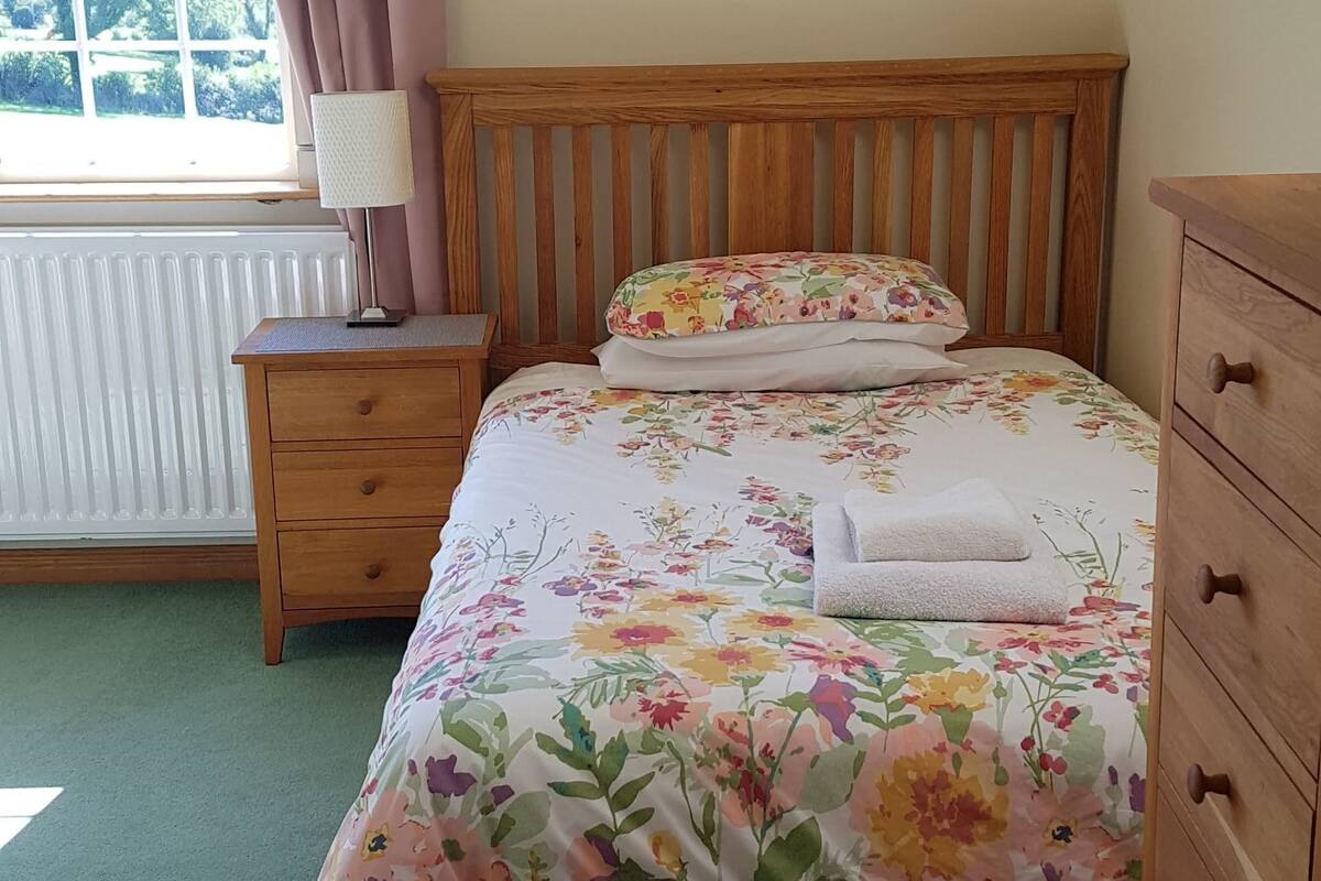 A bedroom with a wooden bed, floral bedding, a nightstand with a lamp, a dresser, and a window with curtains.