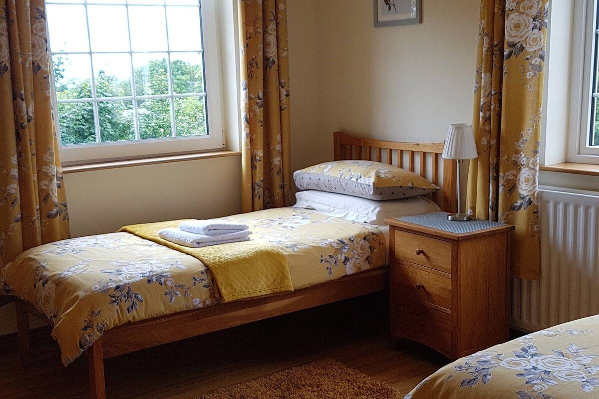 A bedroom with a single bed, a wooden nightstand, a bedside table with a lamp, a window with curtains, and a radiator.