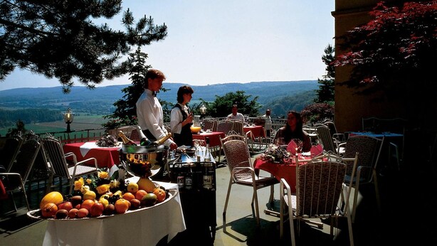 An outdoor dining area with tables set for two, a waiter serving, and a table with a fruit display. - Burghotel Volmarstein (Wetter)