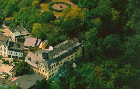 Vista aérea de una zona residencial con varias casas, rodeada de vegetación densa y un parque circular con una fuente.