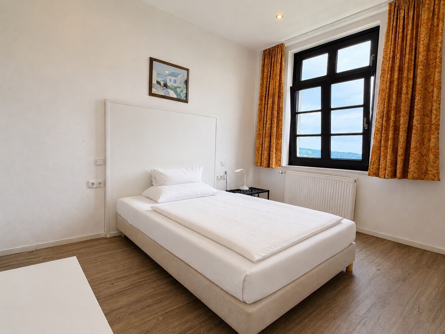A hotel room with a large bed, a bedside table with a lamp, a window with curtains, and a painting on the wall.