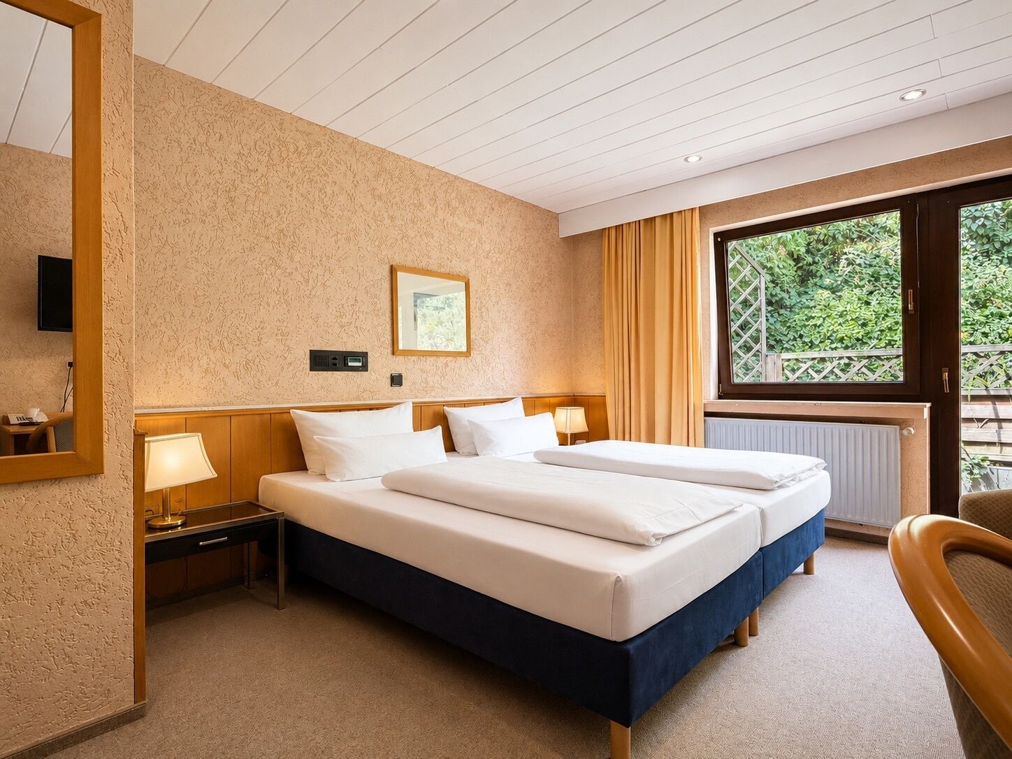 Ein Hotelzimmer mit Doppelbett, Nachttisch mit Lampe, Spiegel und Fenster mit Blick auf Grünflächen.