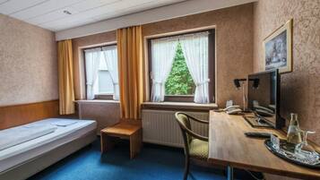 Habitación de hotel con una cama individual, una pequeña mesa de madera, un escritorio con computadora y teléfono, y una ventana con cortinas.