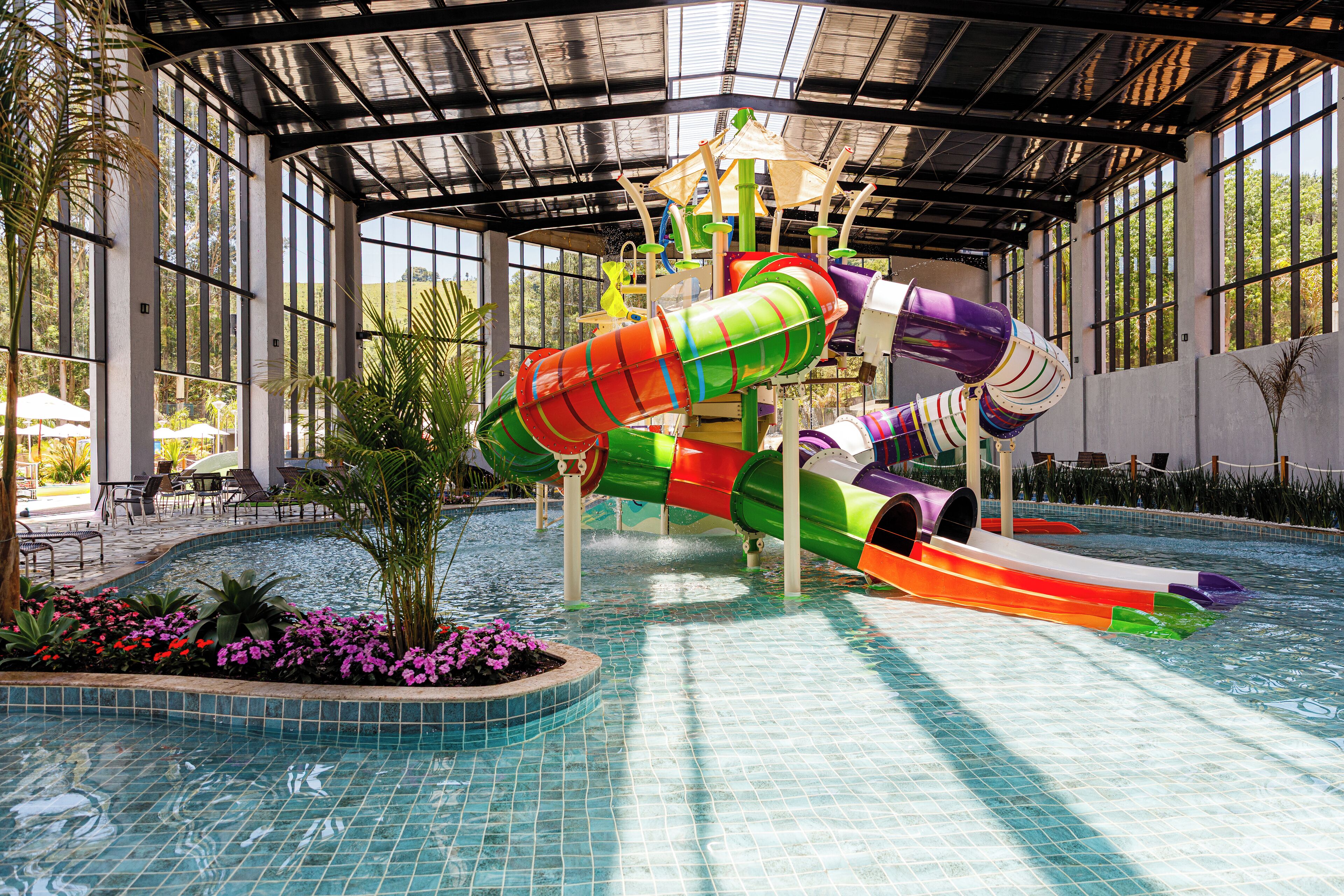 Parque aquático interno com um escorregador de águas colorido e área de piscina com plantas e guarda-sóis.