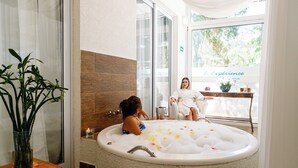 Et spa-værelse med et stort badekar fyldt med bobler, et træbord med en vase blomster og udsigt til grøn bevoksning udenfor.