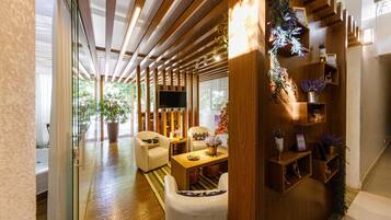 Uma sala de estar moderna com sofá, poltronas, mesa de centro e televisão. Há estantes de madeira com plantas e objetos decorativos.