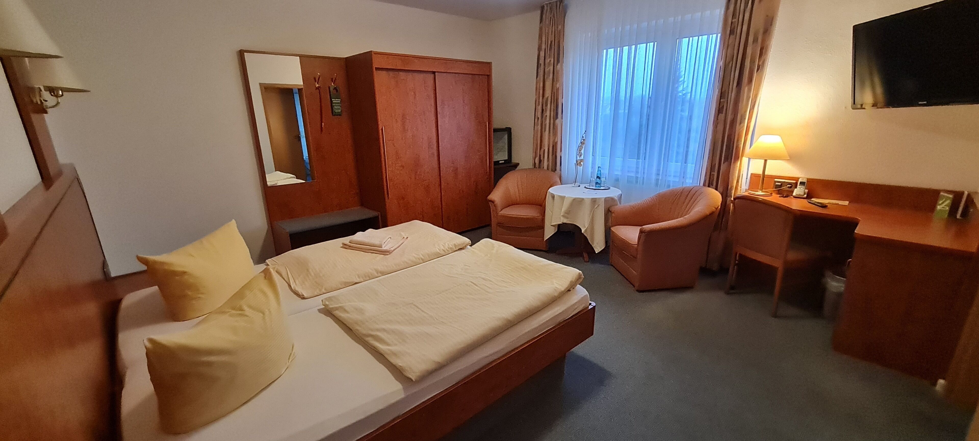 Camera d'albergo con un letto, due poltrone, una scrivania, uno specchio e una finestra con tende.