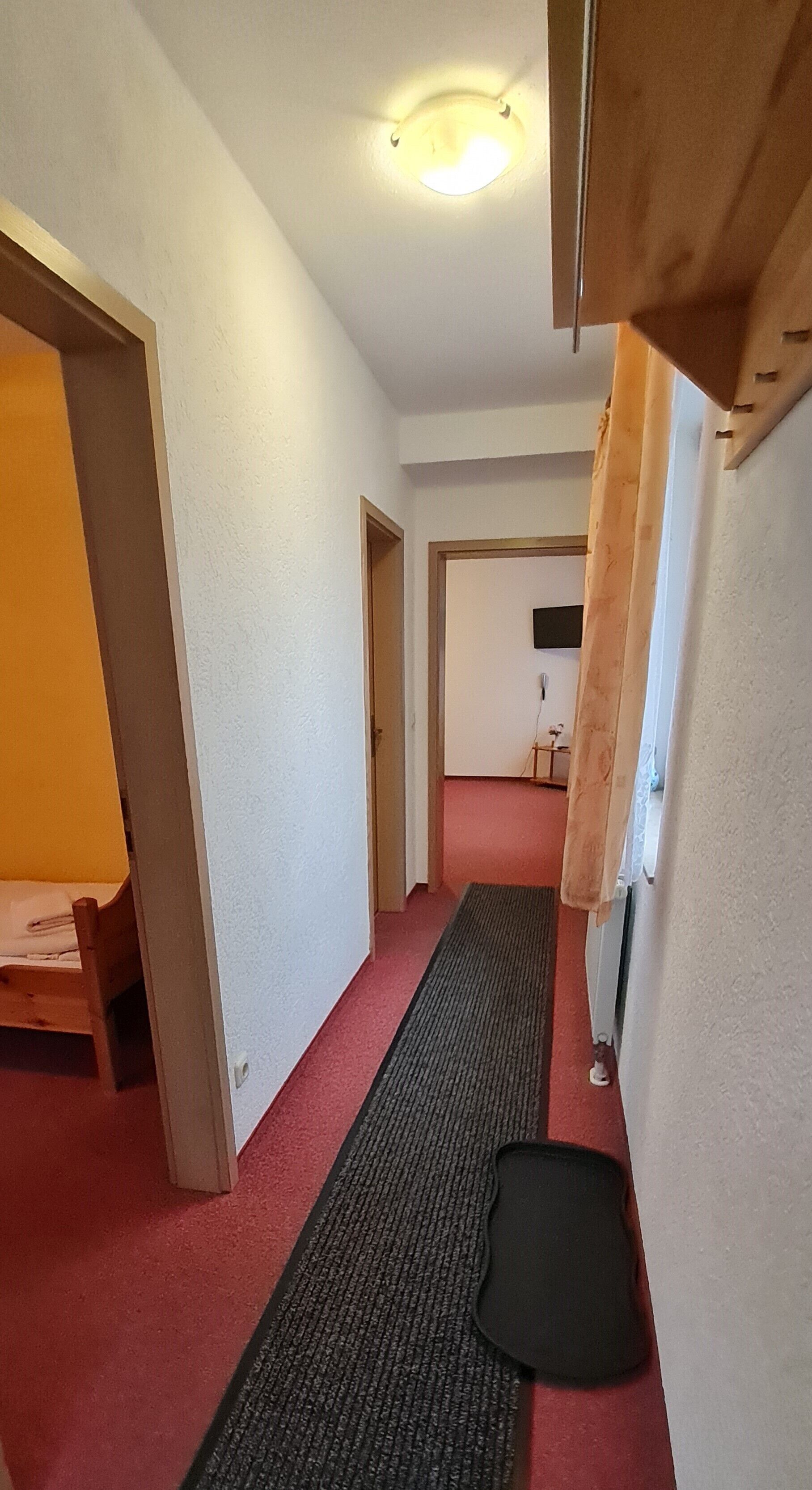 Ein schmaler Flur mit einer hölzernen Bank links, einer schwarzen Matte am Boden und einer Tür am Ende.