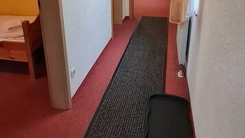 Ein schmaler Flur mit einer hölzernen Bank links, einer schwarzen Matte am Boden und einer Tür am Ende.