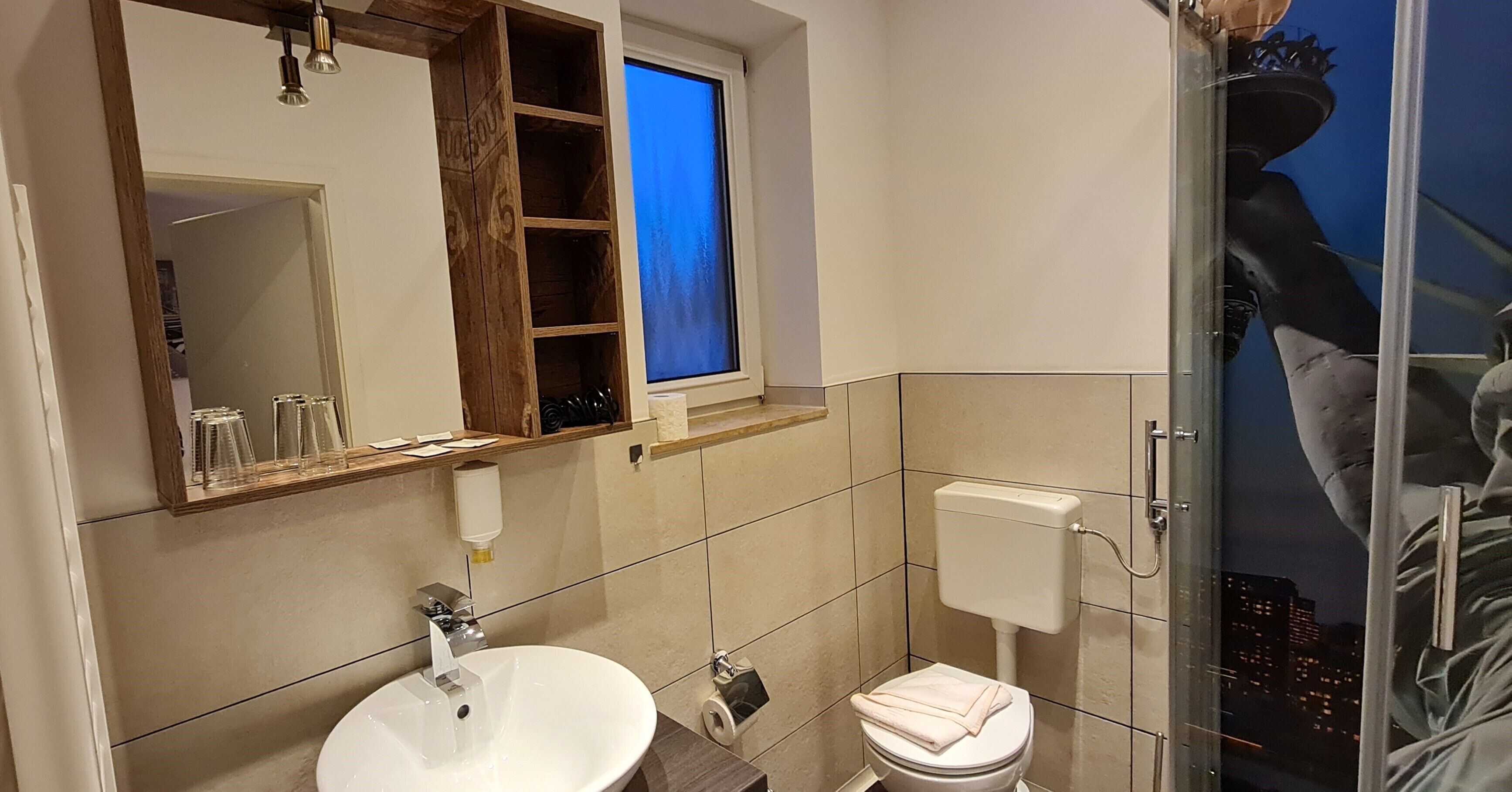 Casa de banho com lavatório branco, sanita e chuveiro. Tem uma janela com vista para a cidade à noite.