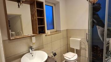 Casa de banho com lavatório branco, sanita e chuveiro. Tem uma janela com vista para a cidade à noite.