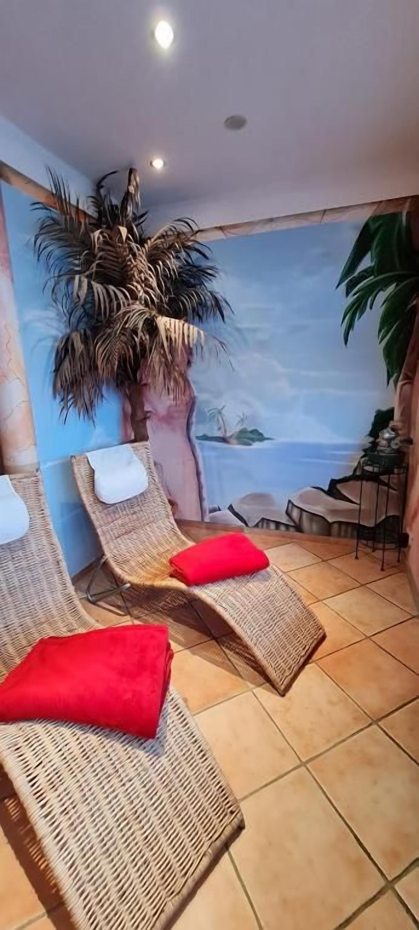 Quarto com poltronas de vime, almofadas vermelhas, um mural com paisagem de praia e uma palmeira.