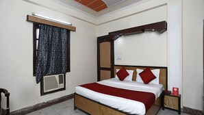 Une chambre bien rangée, avec un lit, une fenêtre avec des rideaux, un climatiseur fixé au mur et une petite table de chevet sur laquelle se trouve un téléphone.