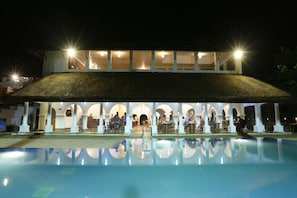 A hotel with a swimming pool, covered outdoor seating area, and a thatched roof structure.