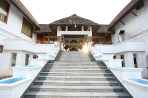 A grand staircase leading to a covered area with a thatched roof, flanked by white buildings with balconies. - Palette Resorts - Le Pondy (Puducherry)