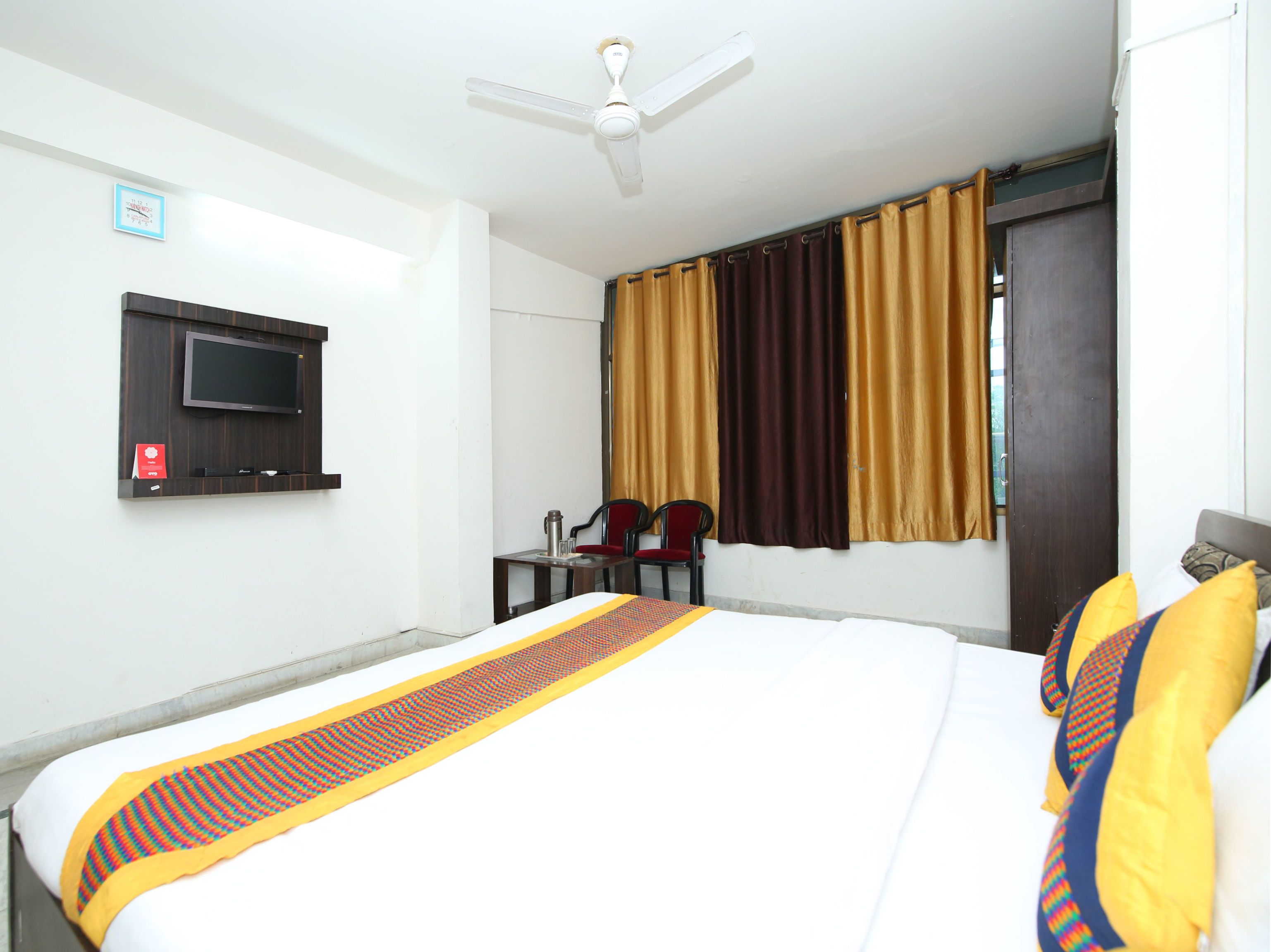 A hotel room with a bed, a TV mounted on the wall, a ceiling fan, and a window with curtains.