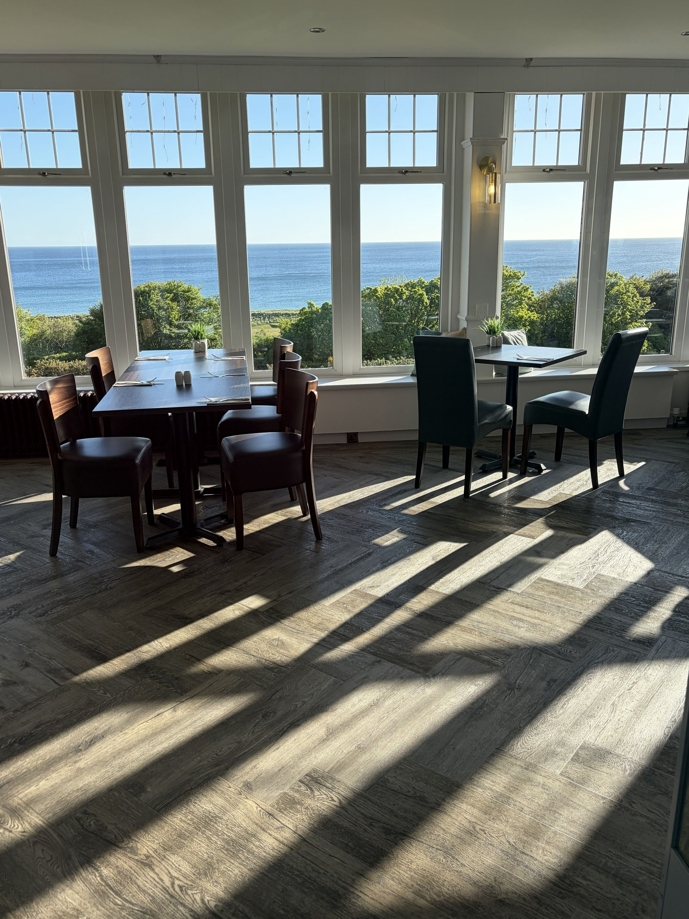 A dining area with a view of the sea, large windows, and wooden flooring.
