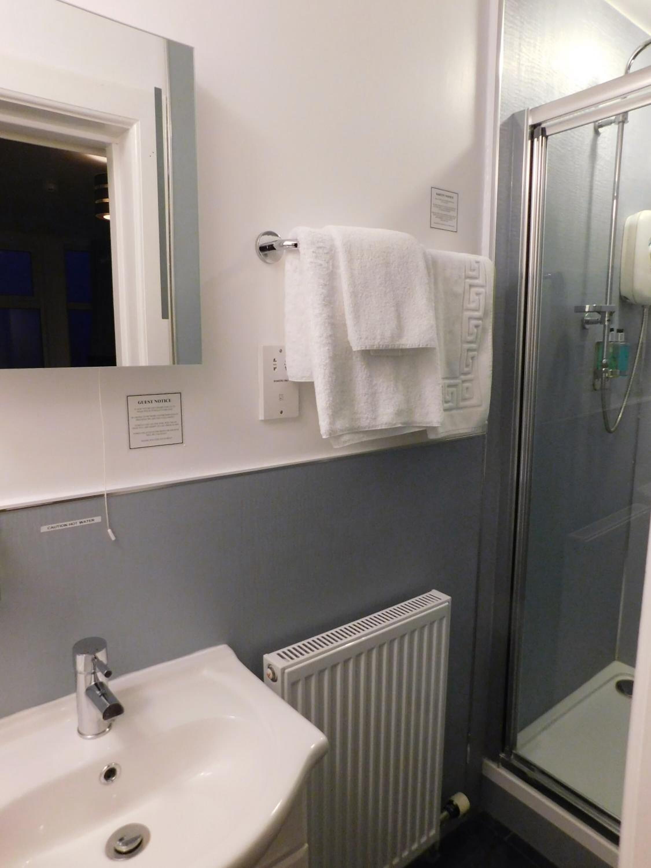 A bathroom with a white sink, a mirror, a towel rack with two towels, a glass shower enclosure, and a caution sign for hot water.