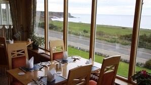 A dining area with a table set for four, large windows offering a view of a coastal road and cliffs. - Cullen Bay Hotel (Buckie)