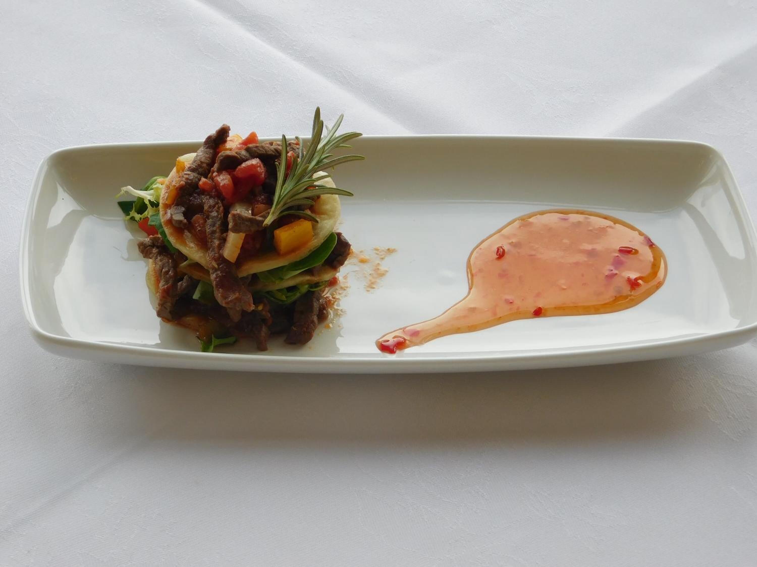 Un plato rectangular blanco con guiso de carne y verduras, adornado con una ramita de romero y una cucharada de salsa al lado.