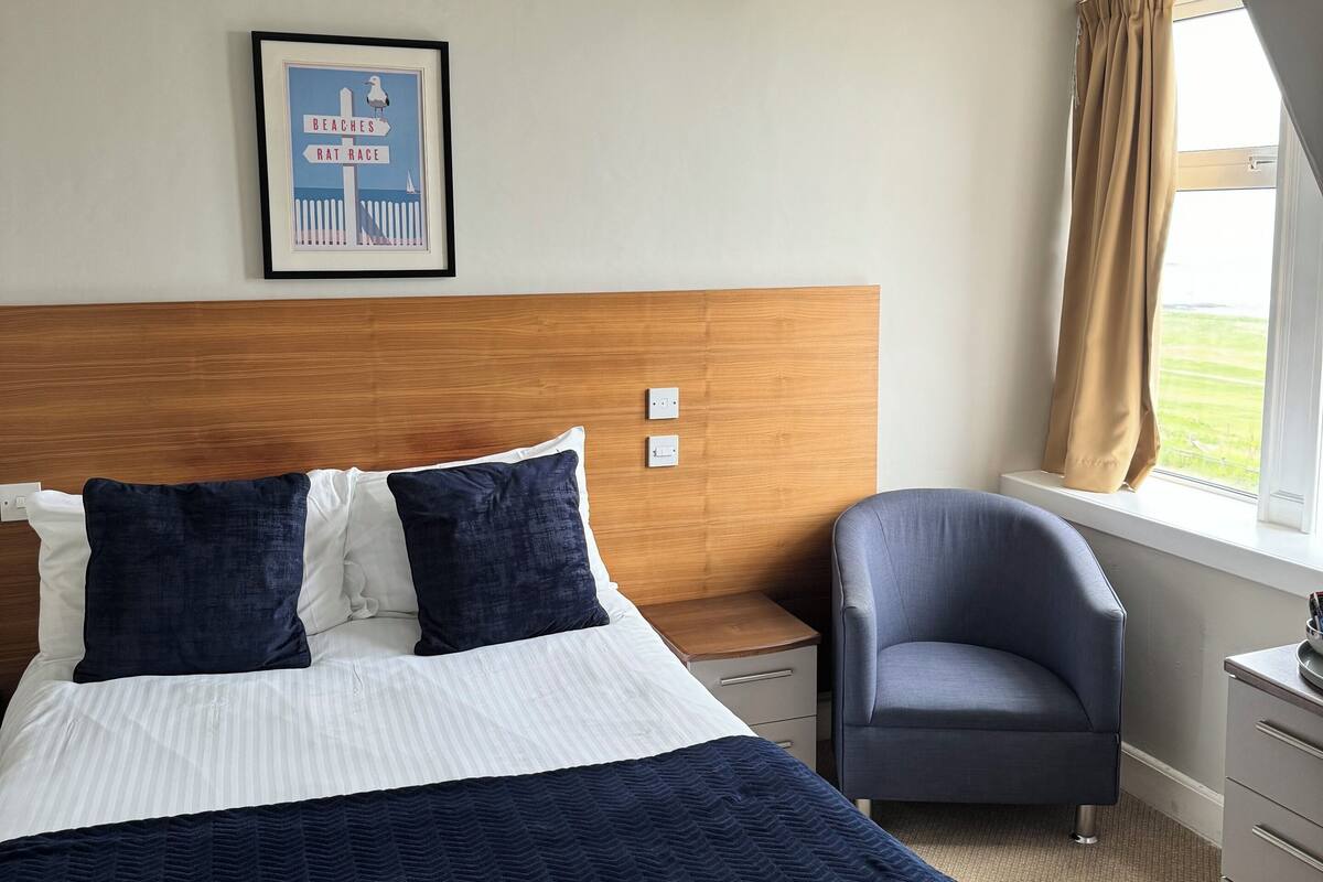 A hotel room with a large bed, a wooden headboard, a framed picture on the wall, a window with curtains, and a desk with a chair.