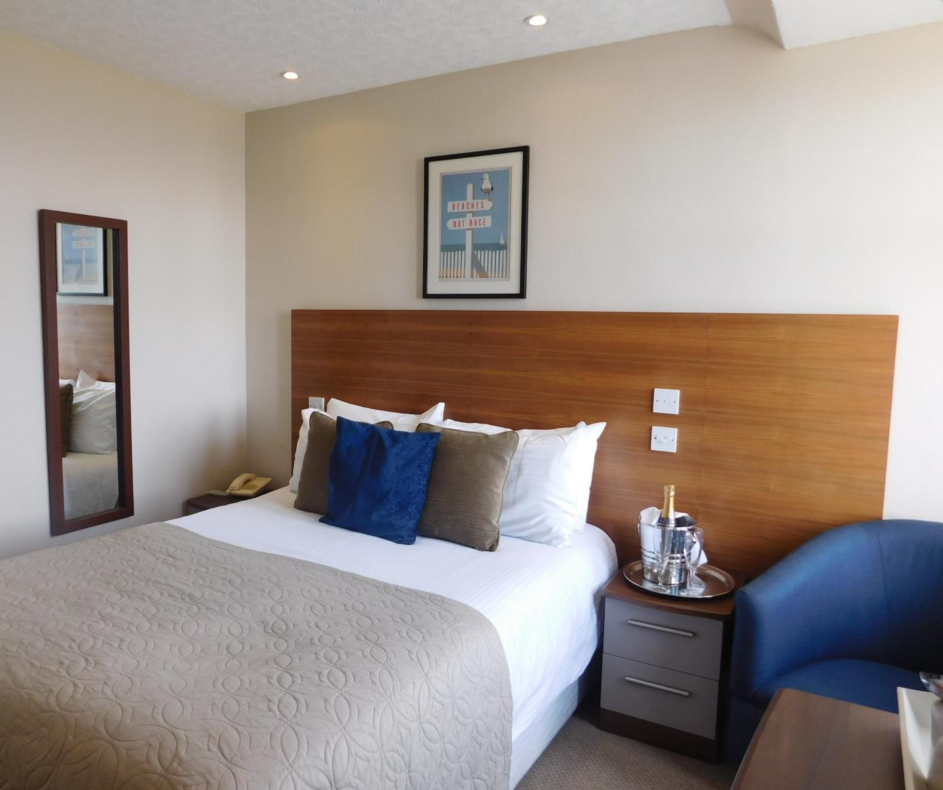A hotel room with a large bed, a wooden headboard, a bedside table with a bottle of champagne, a mirror, and a framed picture on the wall.