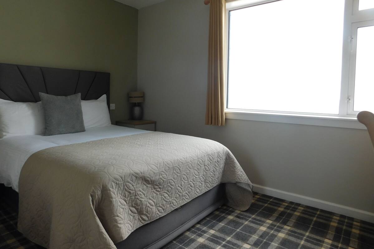 A neatly made bed with a headboard, a bedside table with a lamp, and a window with curtains.