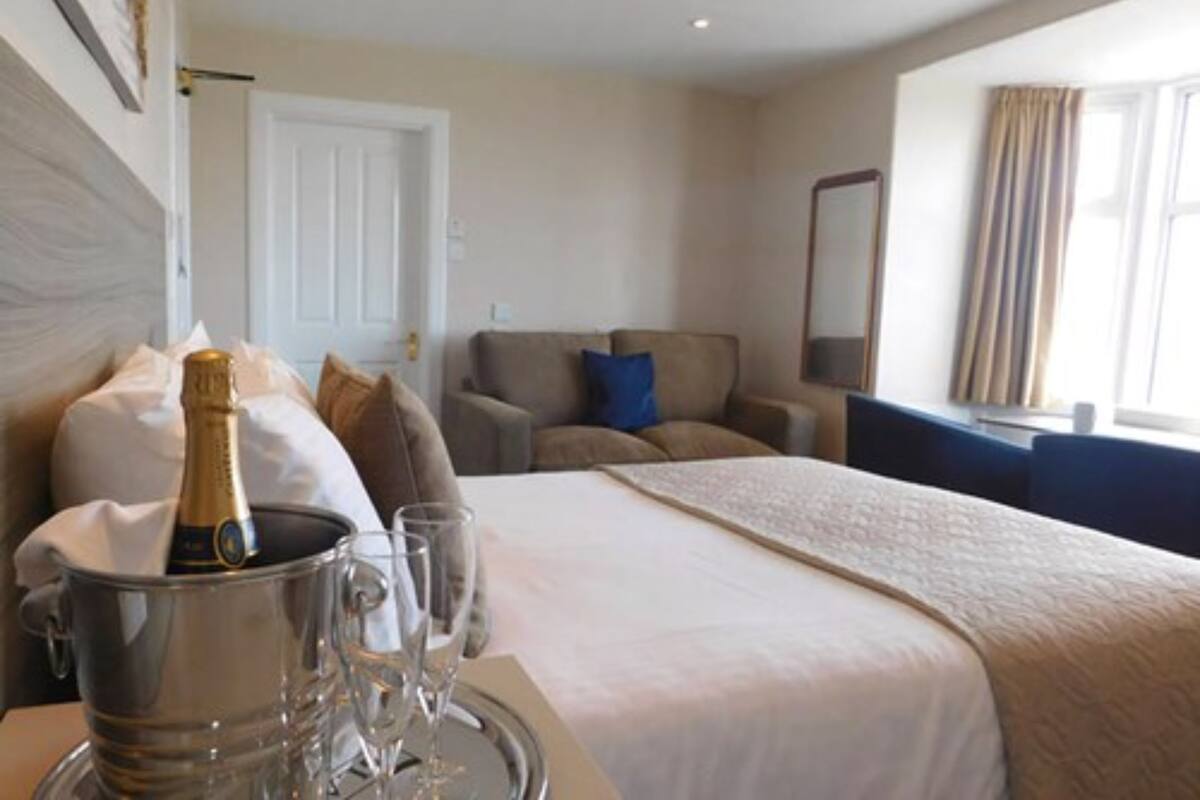A hotel room with a bed, a bedside table with a champagne bottle and glasses, a sofa, a desk, and a window with curtains.
