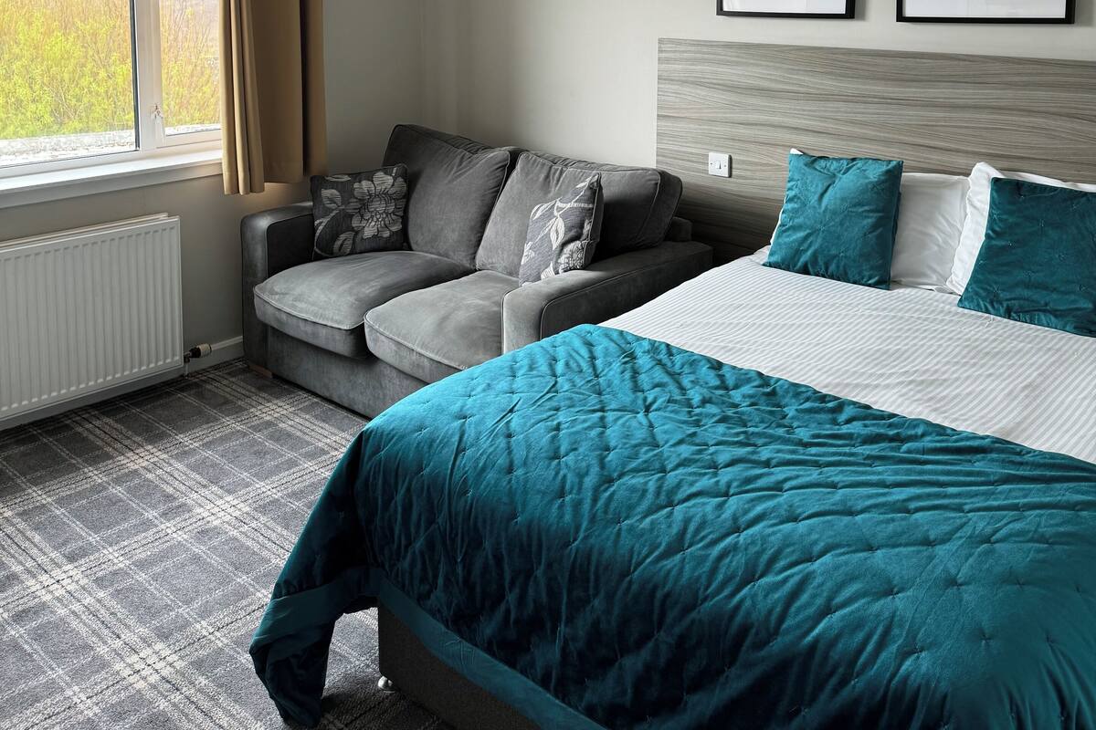 A modern bedroom with a grey sofa, a bed with teal bedding, and two framed black and white photographs on the wall.