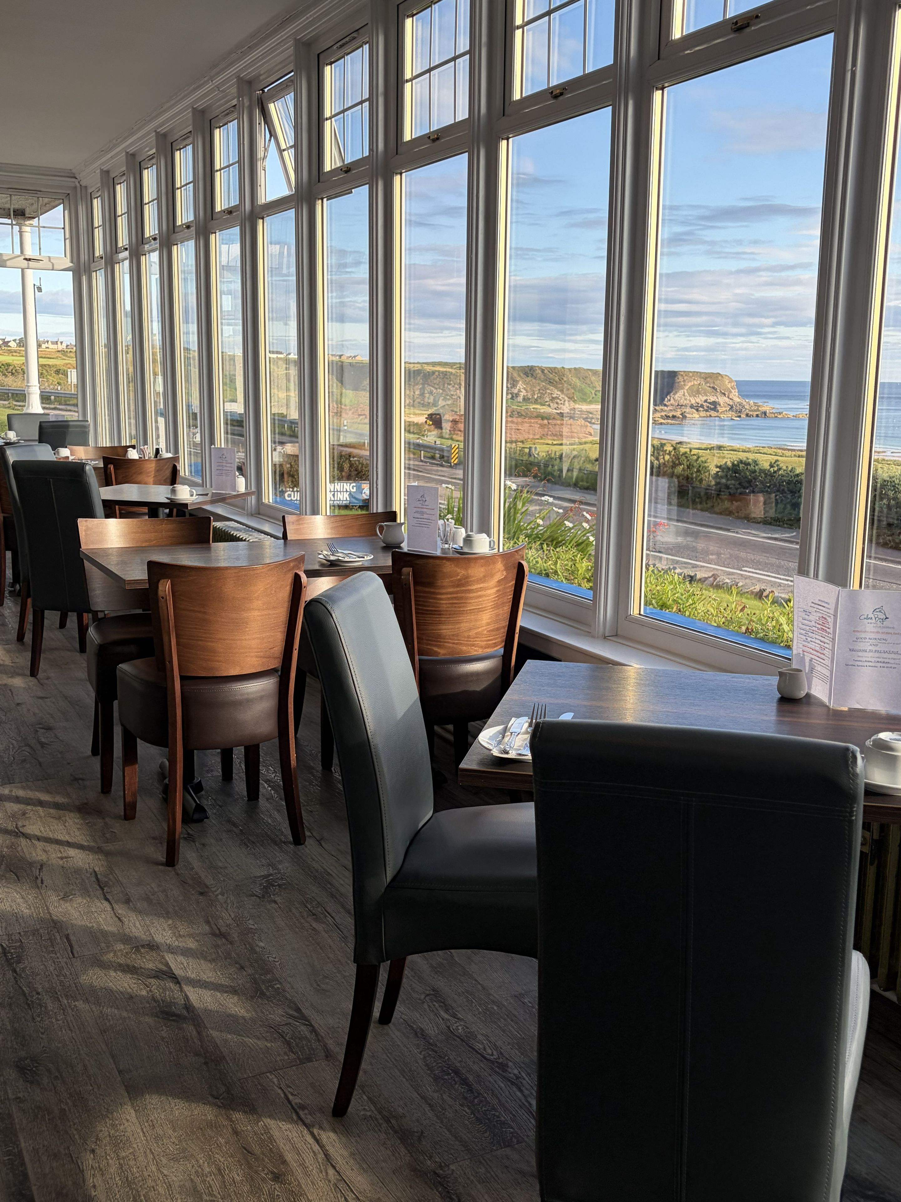 Restaurante com grandes janelas que oferecem vista para a costa. Mesas e cadeiras dispostas para refeições.