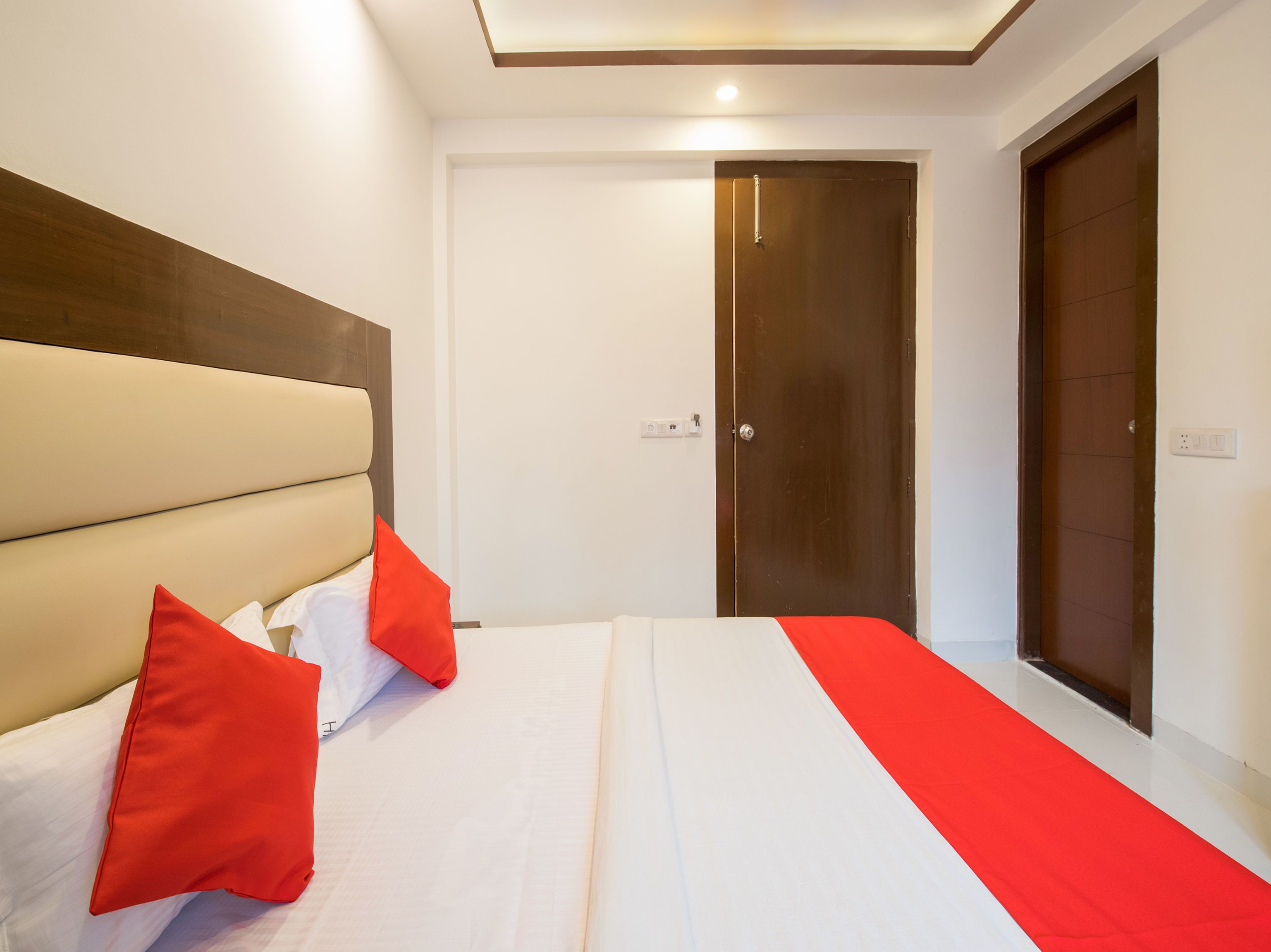 Habitación de hotel con una cama grande, cabecera de madera, almohada decorativa roja, colcha blanca y un tapete rojo.