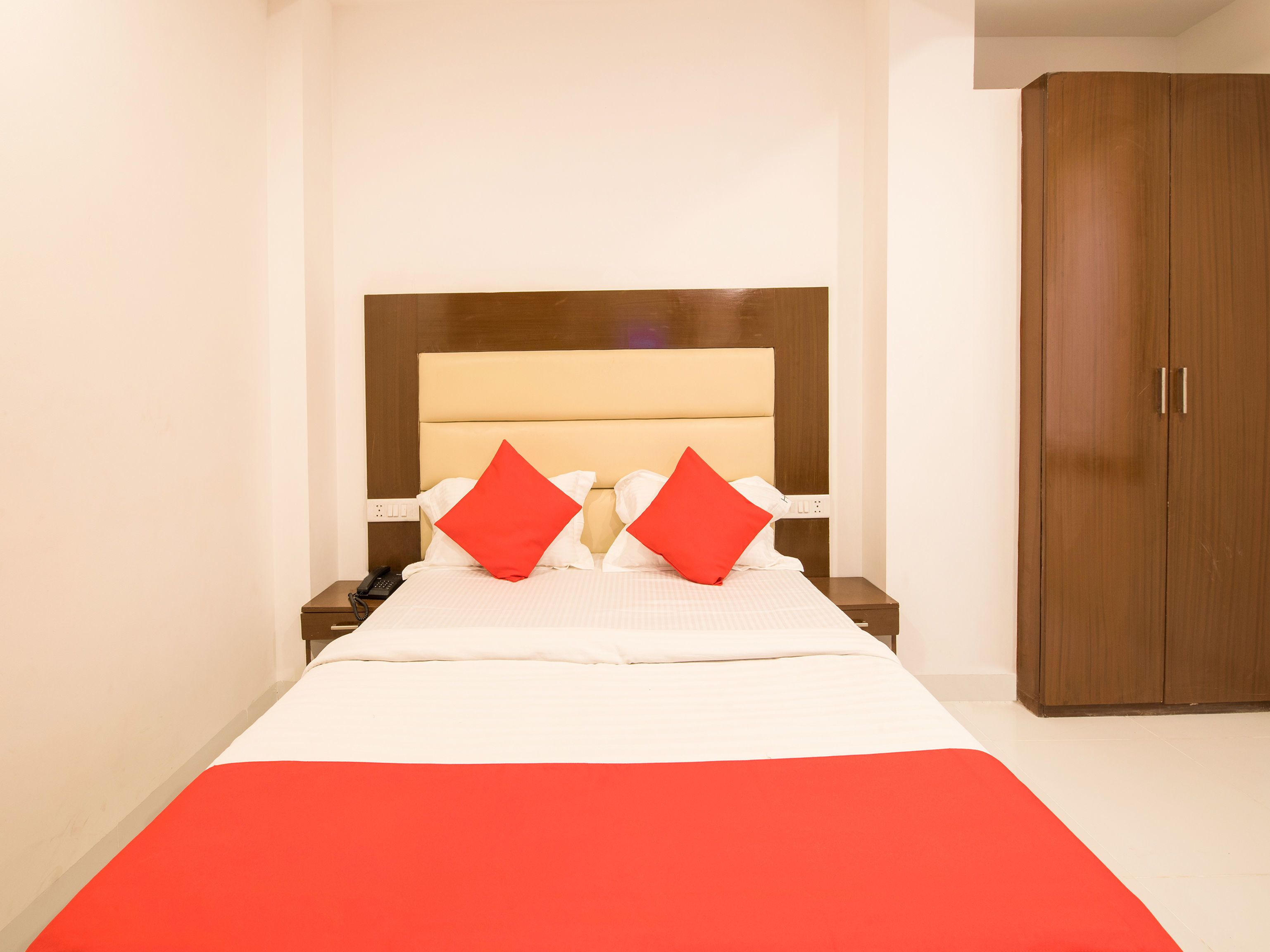 A neatly made bed with a red accent, a wooden wardrobe, and a bedside table with a phone.