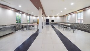 A spacious indoor area with multiple tables and chairs arranged in rows, large windows on the walls, and ceiling fans.