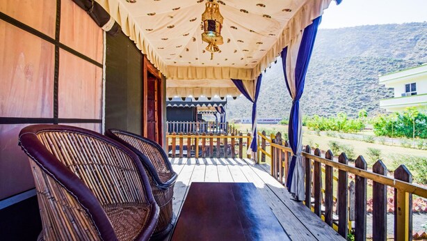 A wooden deck with wicker chairs and a table, overlooking a garden and a mountain.