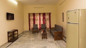 Un espace de vie compact comprenant un canapé, deux fauteuils, une petite table, un réfrigérateur, un ventilateur de plafond et un téléviseur fixé au mur.