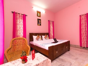 A pink-themed room with a bed, a wicker chair, a small table with a red flower, and a sign.