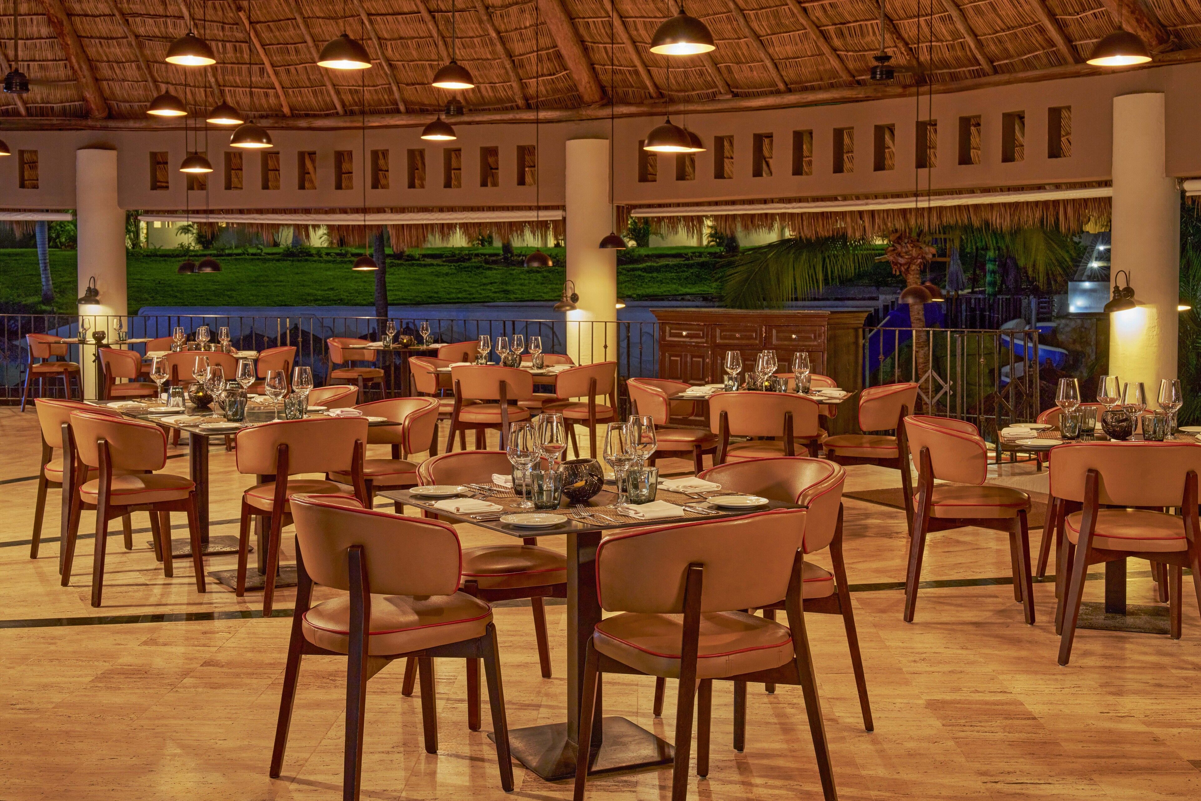 A restaurant with multiple tables and chairs, a thatched roof, and a view of a lush landscape.