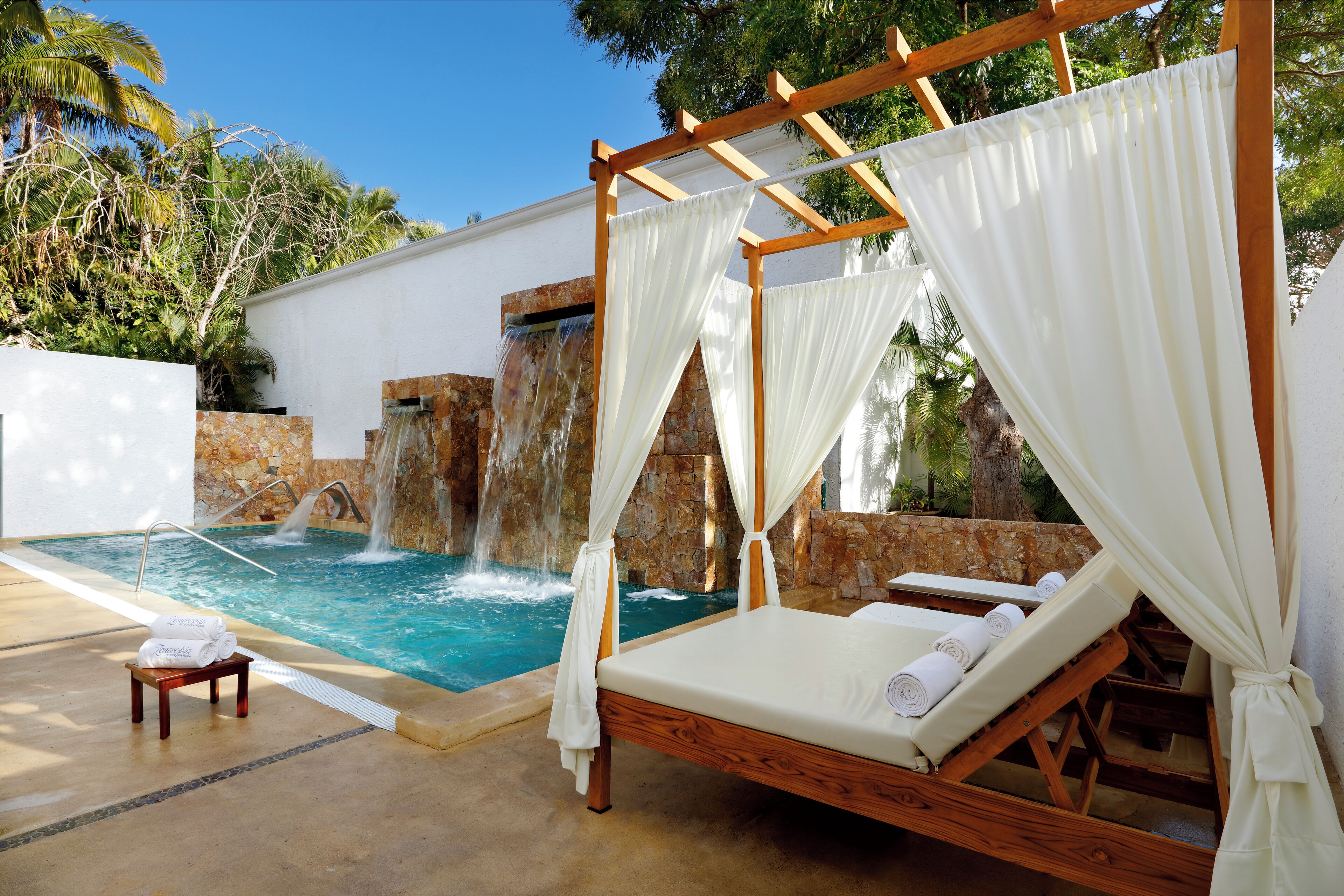 A poolside area with a wooden bed, white curtains, and a small table with towels.