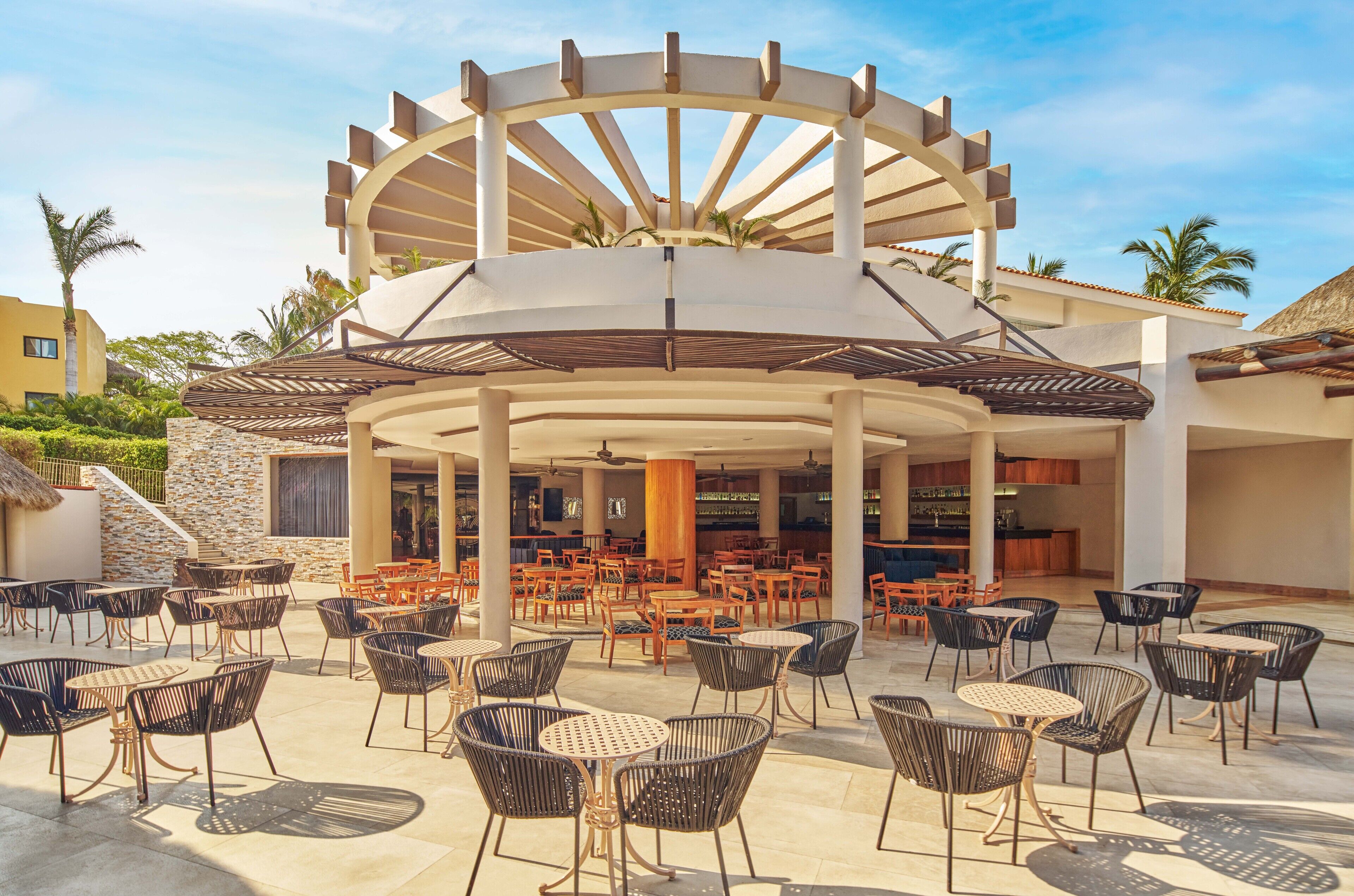 Un patio al aire libre con diversas opciones de mobiliario, incluyendo mesas y sillas redondas, y un elemento arquitectónico central con diseño circular.