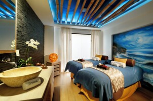 A modern hotel room with a large stone sink, two beds with blue bedding, a large painting of a beach scene, and a bamboo ceiling design.