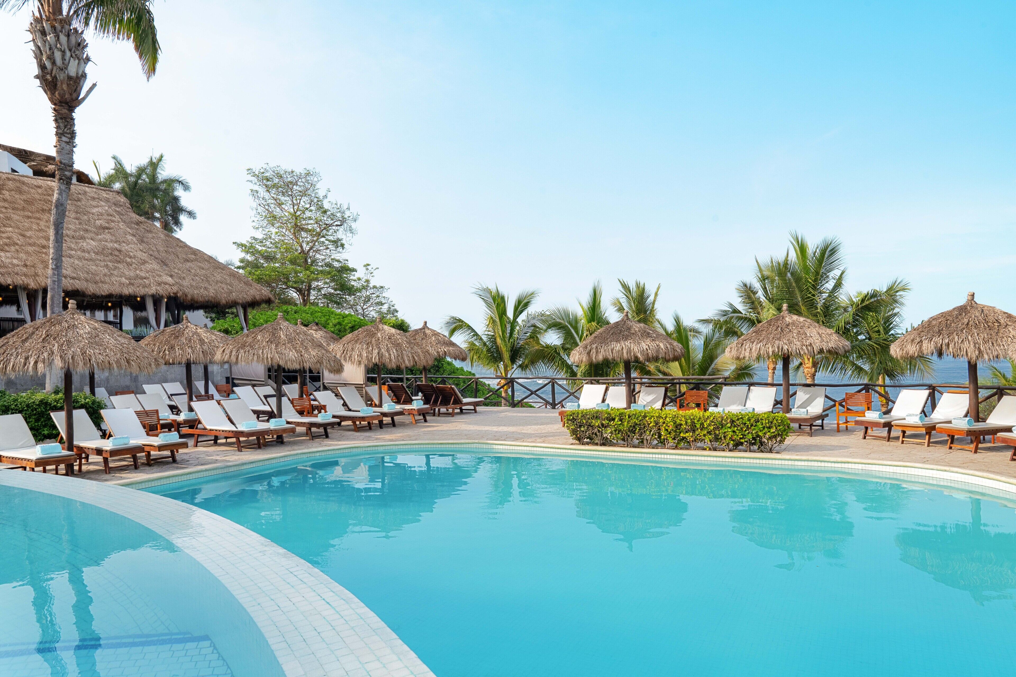 A pool area with lounge chairs and thatched umbrellas, surrounded by palm trees and a clear sky.