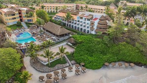 Ein Resort mit Schwimmbad, Strandbereich mit Liegestühlen und mehreren Gebäuden mit Reetdächern.