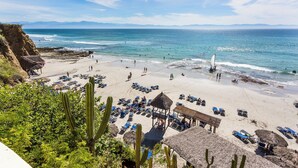 Een strand met rieten hutten, ligbedden en een helder uitzicht op de oceaan.