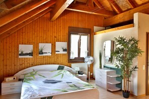 A bedroom with a wooden ceiling, a bed with a green leaf pattern, a bedside table, a mirror, a potted plant, and a window with curtains. - Hôtel du Port (Estavayer-le-Lac)