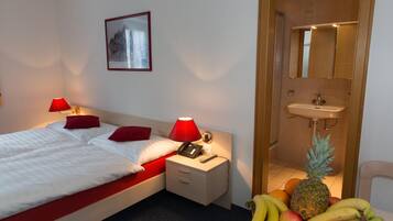 Habitación de hotel con cama, lámparas de noche, mesita de noche, cajón pequeño, teléfono en la pared, cuadro enmarco, baño con lavabo y espejo, y una cesta de frutas en el suelo.