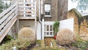 A narrow alleyway with a wooden staircase leading to a white door, surrounded by brick walls and potted plants. - The Chelsea Apartment (London)