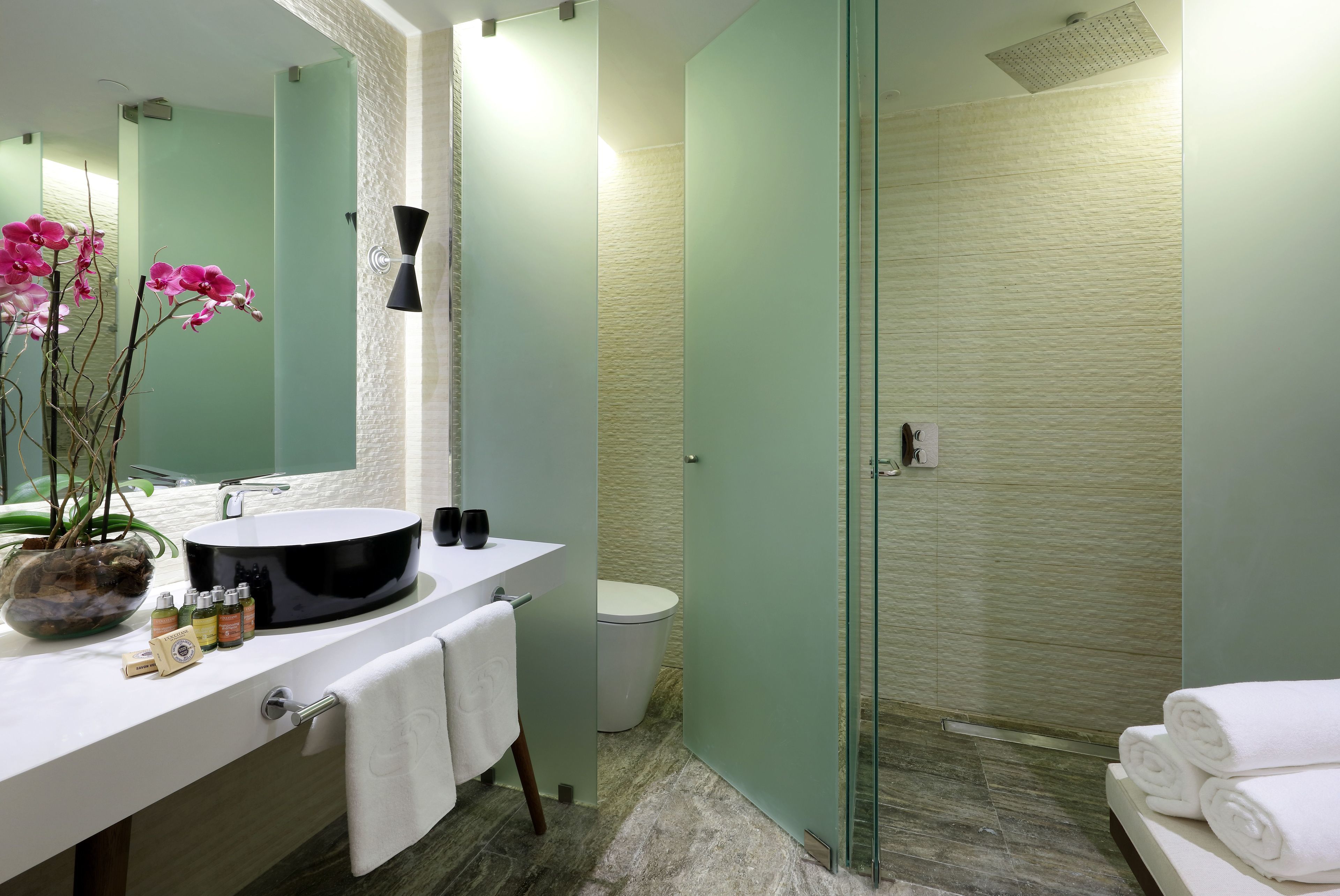 A modern bathroom with a large mirror, a round sink, a glass shower enclosure, and a potted orchid.