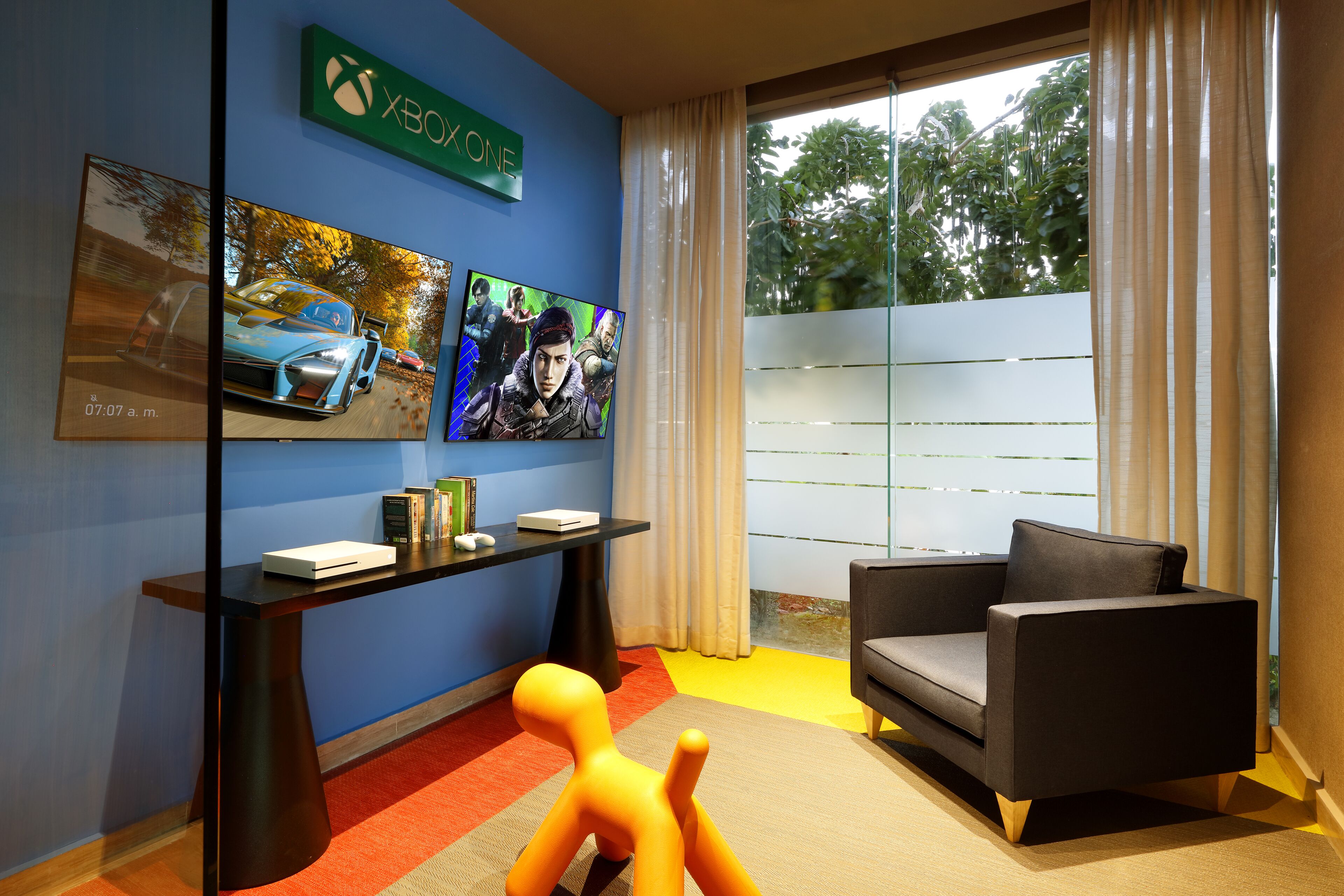 Una habitación moderna con una pared azul decorativa, un ventanal con cortinas, un sofá gris y un mueble bajo con dispositivos electrónicos.