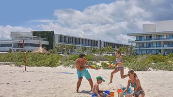 Una playa con gente jugando y construyendo castillos de arena, edificios modernos al fondo y un cielo despejado.