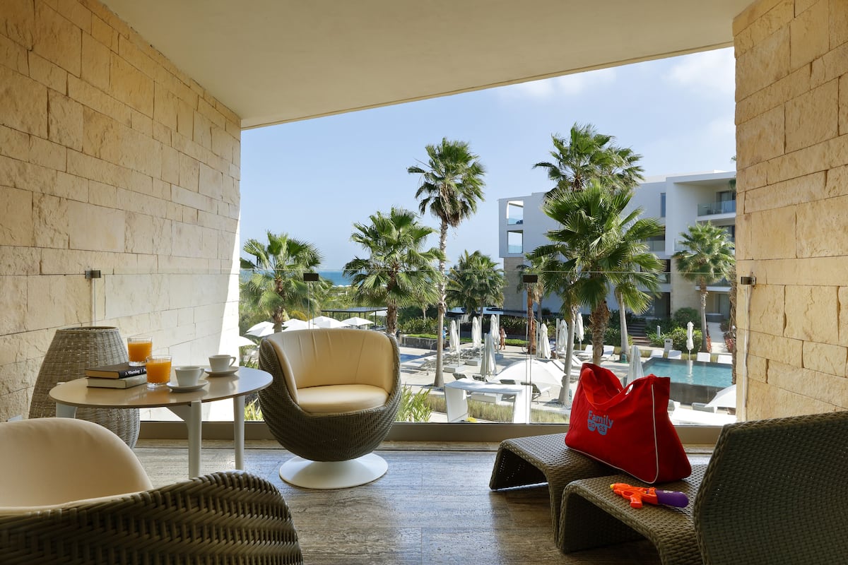 A balcony with wicker furniture, a table with a drink and book, and a view of a pool and palm trees.